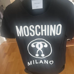 Svart Moschino Milano t-shirt - Svart t-shirt från Moschino med stor vit logga och Milano-tryck på bröstet. Klassisk rund halsringning och korta ärmar. Tillverkad i mjuk bomull för en skön känsla och snygg passform. Perfekt statement-plagg för dig som gillar streetstyle. Storleken är 46 EU och passar S-M. Skicket är bra och sparsamt använt. 