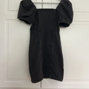 Svart jeansklänning med puffärmar - Cool svart kort jeansklänning med markerade sömmar och puffärmar i stolek 32. Klänningen har en tight passform. Materialet är denim och man kan ha den med ärmarna upp på axlarna eller off shoulder. Aldrig använd