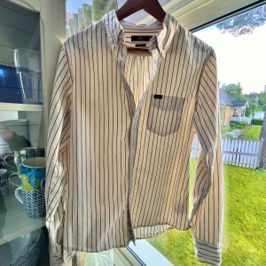 Vit randig skjorta från Lee - Vit skjorta från Lee med tunna svarta vertikala ränder och klassisk krage. Regular fit-modell med lång ärm, bröstficka och knäppning framtill. Skjortan är i mjuk bomull och passar perfekt till jeans eller chinos för en clean look.