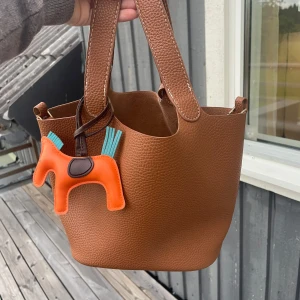 Brun skinnhandväska med hästcharm - Snygg brun handväska i grynigt skinn med vita sömmar och två rejäla handtag. Väskan har en cool orange hästcharm med blå detaljer och ett nyckelhölster i matchande skinn. Perfekt storlek för vardagens essentials och ger en lyxig vibe. Vet inte om det är äkta läder eller ej och vet inte märke, köpt begagnat 😊