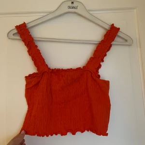 Röd/orange smockad linnetopp Vero Moda - Superfin röd/orange linnetopp från Vero Moda med smockad struktur och volangdetaljer på axelbanden och nederkanten. Toppen är croppad och har breda axelband som ger en söt och trendig look. Perfekt för varma dagar och sommarvibbar!