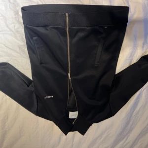 Svart bomberjacka från Les Deux - Snygg svart bomberjacka från Les Deux med klassisk ribbad krage och muddar. Jackan har dragkedja framtill, två fickor och stilren design med diskret logga på bröstet. Perfekt för dig som gillar en clean och modern look.