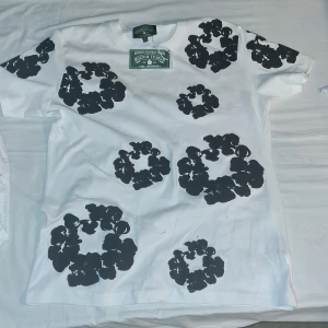 Vit t-shirt med svart blommigt tryck Denim Tears - Säljer en vit t-shirt från Denim Tears med stora svarta blommiga tryck över hela tröjan, både fram och bak. Klassisk passform med rund hals och korta ärmar. Tillverkad i mjuk bomull, perfekt för dig som gillar statement-plagg.