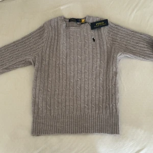 Grå kabelstickad tröja Polo Ralph Lauren - Kabelstickad grå tröja från Polo Ralph Lauren med klassisk rund halsringning och broderad svart logga på bröstet. Tröjan är i mjuk ull och har ribbade muddar vid ärmslut och nederkant. Perfekt för lager-på-lager under kyliga dagar. Helt nytt skick, aldrig användt den förutom när jag har prövat den. Jag säljer denna eftersom att den inte passade mig för den var för liten (jag beställde på nätet). Det är bara att ställa frågor om ni undrar något! 😁