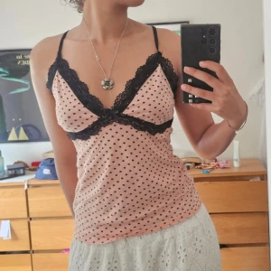 Rosa prickig linnetopp med spets - Supersöt rosa linnetopp med svarta prickar och tunna axelband. Toppen har svart spets längs urringningen och under bysten samt en liten rosett framtill. Perfekt för en trendig och feminin look. Mjuk och skön bomullskänsla.