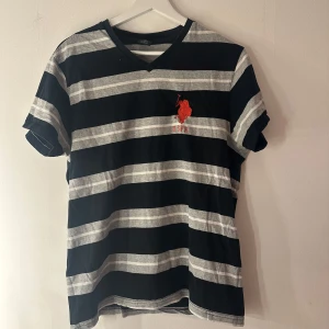 Randig t-shirt från U.S. Polo Assn. - Snygg t-shirt från U.S. Polo Assn. i svart och grått med breda horisontella ränder och röd broderad logga på bröstet. T-shirten har v-ringning och är tillverkad i 100% bomull. Perfekt för en chill och sportig stil. Litet litet hål tyvärr, se bild 4