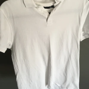 Vit pikétröja från 157, storlek M - Klassisk vit pikétröja från 157 i storlek M. Tröjan har korta ärmar, krage och knäppning med två knappar framtill. Tillverkad i bomull för en skön och luftig känsla. Perfekt till jeans eller shorts för en clean och enkel stil.