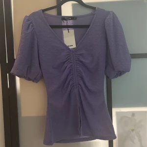 Lila v-ringad topp från Bik Bok - Superfin lila topp från Bik Bok med v-ringning och puffiga korta ärmar. Toppen har snygg rynkdetalj framtill och är gjord i ett strukturerat tyg som ger en cool känsla. Perfekt för dig som vill sticka ut lite extra med en trendig färg och form.