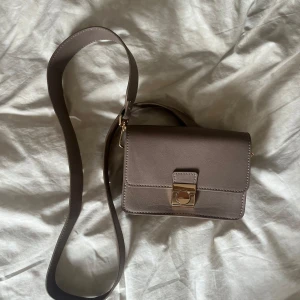 Brun axelväska med guldspänne - Snygg brun axelväska i skinn med stilren, fyrkantig form och bred axelrem. Väskan har ett elegant guldigt spänne framtill och fack inuti för smart organisering. Perfekt storlek för det viktigaste och passar till många outfits.