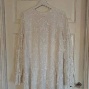 Vit blommig klänning från NA-KD - Superfin vit klänning från NA-KD med broderade blommor, långärmad och med snygg öppen rygg och knytband. Klänningen har en luftig passform och volangdetaljer nertill. Perfekt för sommarens alla tillfällen!