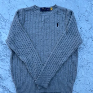 Grå kabelstickad tröja Polo Ralph Lauren - Säljer en stilren nyskick kabelstickad grå tröja från Polo Ralph Lauren med klassisk rund hals och broderad svart logga på bröstet i storlek M. Jag köpte dem nyligen av en annan person men säljer dem vidare då de tyvärr är för små för mig. 