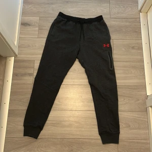 Svarta mjukisbyxor Under Armour M - Svarta mjukisbyxor från Under Armour med röd logga framtill och vit text på benet. Byxorna har snörning i midjan, ribbade muddar och en dragkedjeficka på sidan. Sköna och loose fit, perfekta för chill eller träning.
