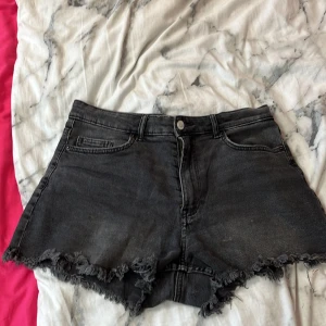 Svarta jeansshorts med fransig kant - Snygga svarta jeansshorts med rå, fransig kant och klassisk femficksdesign. Shortsen har en hög midja och dragkedja med knapp framtill. Perfekta för sommardagar när du vill ha en cool och avslappnad look.