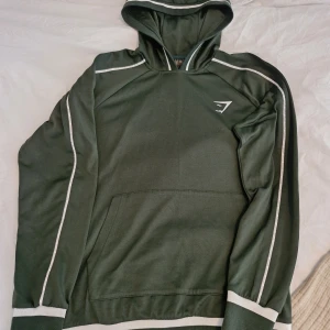 Grön hoodie från Gymshark med vita detaljer - Mörkgrön hoodie från Gymshark i storlek S med vita ränder längs ärmar och huva. Tröjan har en klassisk känguruficka framtill och Gymshark-logga på bröstet. Perfekt för träning eller chill, med mjukt material och sportig vibe.
