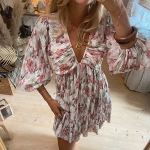 Blommig kortklänning med v-ringning - Superfin kortklänning med blommigt mönster i rosa och vita toner. Klänningen har v-ringning, volangkant nertill och vida, trekvartsärmar. Materialet är lätt och luftigt, perfekt för varma dagar. Snygg passform med markerad midja och plisserad kjol.