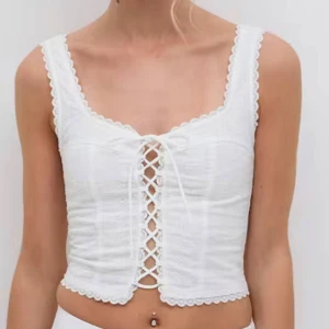 Vit korsett-topp Gina  - Superfin vit korsett-topp med snörning framtill och spetskant runt halsringning och nederkant. Croppad modell med breda axelband och romantisk vibe, perfekt till jeans eller kjol. Tunn och luftig, passar perfekt till varma dagar. Ger rabatt vid snabb affär