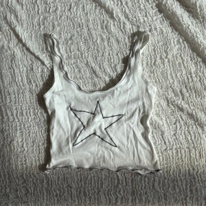Vit croppad linnetopp med stjärna - Superfin vit croppad linnetopp med svart stjärna sydd på framsidan. Toppens kanter har dekorativa svarta sömmar och den har smala axelband. Perfekt för dig som gillar en enkel men ändå cool look.