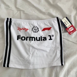 Vit Formula 1 bandeau topp - Snygg vit bandeau topp från Formula 1 med svarta ränder på sidorna och tryck med F1-logga och texten 'Formula 1 Racing' framtill. Topp i mjuk bomullsblandning med lite stretch för perfekt passform. Perfekt för dig som älskar motorsport och streetstyle.