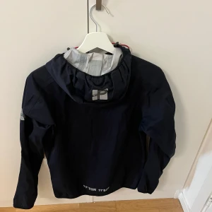 Marinblå vindjacka från Pelle P XS - Snygg marinblå vindjacka från Pelle P med huva, röda snören och dragkedja framtill. Jackan har reflekterande logga på ryggen och ärmarna samt praktiska fickor med dragkedja. Perfekt för blåsiga dagar och outdoor vibes.