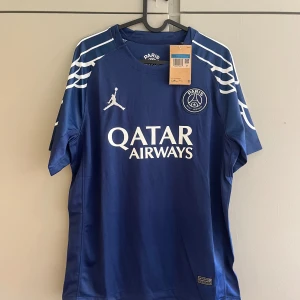PSG blå matchtröja Jordan x Nike - Paris Saint-Germain fotbollströja i mörkblått med vita detaljer och tryck. Klassisk kortärmad modell med klubbmärke, Jordan-logga och Qatar Airways sponsortryck på bröstet. Tillverkad i lätt och ventilerande polyester, perfekt för träning eller match.