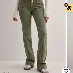 Olivgröna bootcut jeansbyxor - Snygga olivgröna jeansbyxor från Wrangler med bootcut-snitt och hög midja. Byxorna har klassiska fem fickor, dubbla knappar i midjan och dekorativa sömmar på bakfickorna. Perfekta för en trendig och avslappnad look.