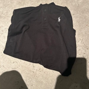 Svart pikétröja från Polo Ralph Lauren - Klassisk svart pikétröja från Polo Ralph Lauren med vit broderad logga på bröstet. Tröjan har korta ärmar, krage och knäppning med tre knappar. Tillverkad i mjuk bomull som känns skön mot huden. Perfekt för en clean och stilren look.