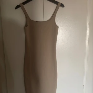 Klänning - En stilren beige kortklänning med smala axelband och figurnära passform. Klänningen är ärmlös och har en enkel, minimalistisk design som passar perfekt för en trendig look. Perfekt att styla med sneakers eller klackar.