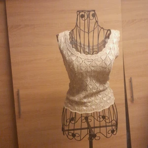 Beige virkad linnetopp med spetskänsla - Supersöt beige linnetopp med virkat mönster och breda axelband. Toppen har en snygg spetskänsla och är perfekt för varma dagar. Den är figurnära och har ett unikt, handgjort utseende som sticker ut.