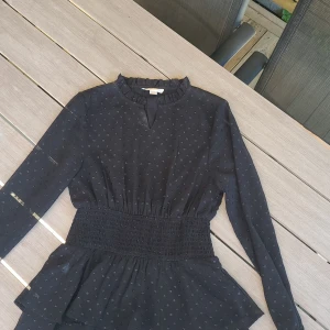 Svart prickig klänning Gina Tricot - Supersöt svart klänning från Gina Tricot med små prickar, långärmad och smockad midja. Volangdetalj nedtill och lätt rynkad krage. Stl 145-152