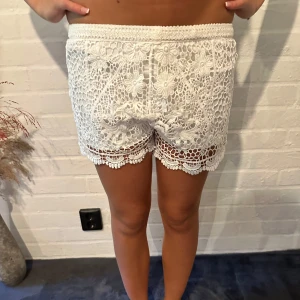 Vita virkade shorts med spetsdetaljer - Superfina vita shorts med virkad look och blommiga spetsdetaljer. De har elastisk midja och snyggt mönstrad kant nertill. Perfekta för sommaren när du vill ha en chill och söt stil. Lätta och luftiga, passar perfekt till ett linne eller crop top.