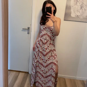 Vit långklänning med röda blommor - Superfin långklänning med smala axelband och v-ringning. Klänningen är vit med rött blommigt mönster och har en hög slits på sidan. Lätt och flowy modell, perfekt för varma dagar. Sitter snyggt i midjan och har öppen rygg.