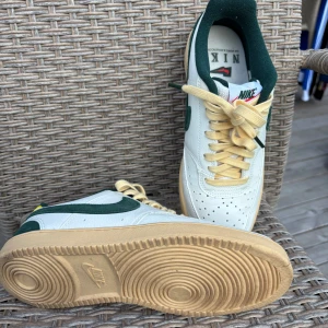 Nike Air Force 1 i vitt och grönt - Snygga Nike Air Force 1 sneakers i vitt skinn med gröna detaljer och beige snörning. Klassisk siluett med perforerad tå och gummisula i naturfärg. Perfekta för dig som gillar retrostil och vill sticka ut med unika färgkombinationer.