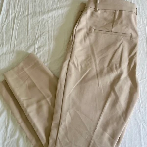 Beige kostymbyxor från H&M - Snygga beige kostymbyxor från H&M i storlek 42. Byxorna har raka ben. Materialet är en mix av bomull, polyester och elastan för en bekväm passform. Aldrig använda!☀️Små i storlek, som en 38!🌸