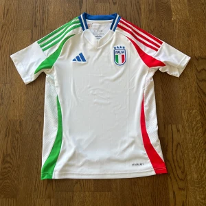 Italien landslag bortatröja 24/25 - En helt ny Italiensk landslagströja från Adidas, storlek 11-12 år. Den är vit med gröna och röda detaljer på sidor och axlar, samt blå krage. Broderad Italien-logga och Adidas-märke på bröstet. Pris gårnatt diskutera.