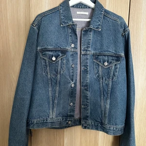 Blå jeansjacka från Our Legacy - Säljer en klassisk blå jeansjacka från Our Legacy. Jackan har raka linjer, två bröstfickor med knappar och snygga sömdetaljer. Tillverkad i robust denim med silverfärgade knappar där varumärket är ingraverat. Perfekt för dig som gillar en tidlös och clean look. Croppad passform.