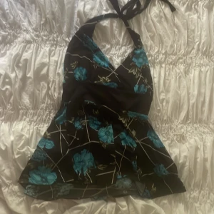 Svart blommig halterneckklänning FMX - Supersöt svart kortklänning från FMX med blått blommönster och halterneck med knyt i nacken. Klänningen har omlott-v-ringning och är gjord i ett stretchigt, mjukt material som sitter snyggt. Perfekt för dig som vill sticka ut med färg och mönster! Man kan använda den som klänning eller topp