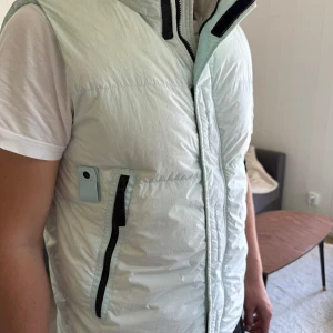 Ljusblå dunväst från Stone Island - Snygg ljusblå dunväst från Stone Island med hög krage, svarta dragkedjor och ikonisk logotyp på sidan. Västen har praktiska fickor med dragkedja och knappdetaljer. Perfekt för lager-på-lager och streetwear-vibe.