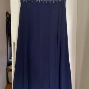 Marinblå långklänning med spets - Elegant marinblå långklänning med spetsliv och vackra pärlbroderier i midjan. Klänningen är ärmlös och har en lätt v-ringning i ryggen. Kjolen är flowy och materialet känns lyxigt, perfekt för bal eller fest. Dragkedja bak för enkel påklädning. Säljs nu vidare för att den inte längre används. Storleken på klänningen är XXL, men den är insydd och upskattas vara mer som XL/L beroende kroppsform. IInga större defekter förutom lite nopprig byst men inget som syns mycket. Väl omhändertagen.