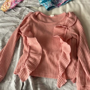 Rosa volangtröja från SHEIN - Supersöt rosa stickad tröja från SHEIN med ribbad struktur och stora volangdetaljer längs ärmar och framsida. Långärmad modell i mjukt material som ger en trendig och lekfull look. Perfekt för dig som gillar att sticka ut lite extra!