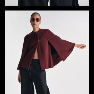 Vinröd cape från Zara i ullmix - Säljer en vinröd cape från Zara med snygga metallspännen framtill. Jackan har vid, svepande passform och är tillverkad i ullmix. Perfekt för dig som vill sticka ut med en elegant och trendig look i höst och vinter.
