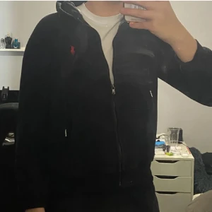 Svart hoodie från Ralph Lauren - Svart hoodie från Ralph Lauren med dragkedja, huva och snörning. Klassisk röd logga broderad på bröstet och stora fickor framtill. Skön passform och perfekt för lager-på-lager. Tillverkad i mjuk bomull.