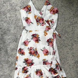 Vit omlottklänning med blommor - Superfin vit omlottklänning med blommigt mönster i rosa, rött och gult. Klänningen har volang nertill och knyt i midjan. Perfekt för sommarens alla tillfällen och ger en fräsch och romantisk vibe.