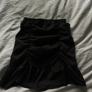 Svart volangkjol från Gina Tricot - Supersnygg svart kortkjol från Gina Tricot med volangkant och rynkade detaljer framtill. Kjolen har en tight passform och en resor i midjan, perfekt för dig som gillar trendiga och feminina plagg. Elastisk midja för extra komfort.