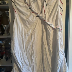 Vit omlottklänning med ränder - Snygg vit kortklänning från Polarn O. Pyret med omlott-skärning och ärmlös design. Klänningen har tunna diagonala ränder i rött och marinblått som ger en sportig vibe. Tillverkad i mjuk bomull, perfekt för varma dagar.