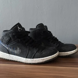 Nike Air Jordan 1 Crater svart/grå - Nike Air Jordan 1 Crater i svart canvas med gråmelerad swoosh och färgstänk på sulan. Skorna har blå zigzag-sömmar och klassisk Jordan-logga på sidan. Hög modell med snörning och återvunnet material i yttersulan. Perfekt för dig som gillar unika detaljer.