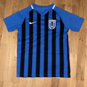Äkta IK Sirius signerad matchtröja Vecchia #22 Nike  - Säljer en blå och svart randig IK Sirius fotbollströja från Nike med klubbmärke på bröstet och tryck på ryggen: Vecchia 22. Tröjan är kortärmad, tillverkad i lätt och ventilerande material. Nummer och namn är tryckta i vitt, och tröjan har en autograf på siffran.