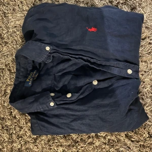 Mörkblå linneskjorta Polo Ralph Lauren storlek L men sitter som xs eller S - Snygg mörkblå skjorta från Polo Ralph Lauren i linne. Klassisk button-down med vita knappar och röd broderad logga på bröstet. Skjortan har lång ärm och avslappnad passform, perfekt till jeans eller chinos.