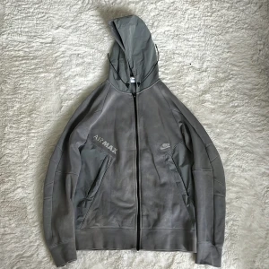 Nike Air Max grå vindjacka med huva - Snygg grå Nike Air Max vindjacka med huva och dragkedja. Jackan har stilrena detaljer, två fickor framtill och diskret Air Max-tryck på bröstet. Perfekt för dig som gillar sportig och clean streetstyle.