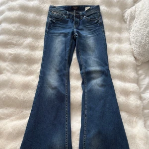 Blå bootcut jeans med strassdetaljer - Snygga blå bootcut jeans från Lola med coola strassdetaljer på bakfickorna. Jeansen har klassisk femficksdesign, normal passform och är tillverkade i mjukt denimtyg. Perfekta för dig som gillar en retroinspirerad look med extra bling.