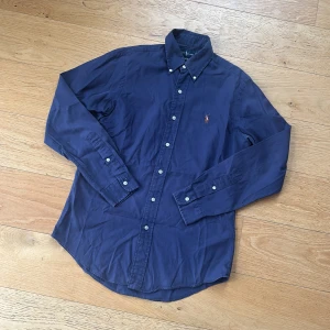 Ralph Lauren skjorta (pris diskuterbart) - !! NY SKICK !!Klassisk mörkblå skjorta från Ralph Lauren i custom fit (SLIMFIT) bomull, custom fit. Skjortan har knappar hela vägen, button down-krage och den ikoniska broderade polospelaren på bröstet. Perfekt för dig som gillar stilrena och tidlösa plagg.